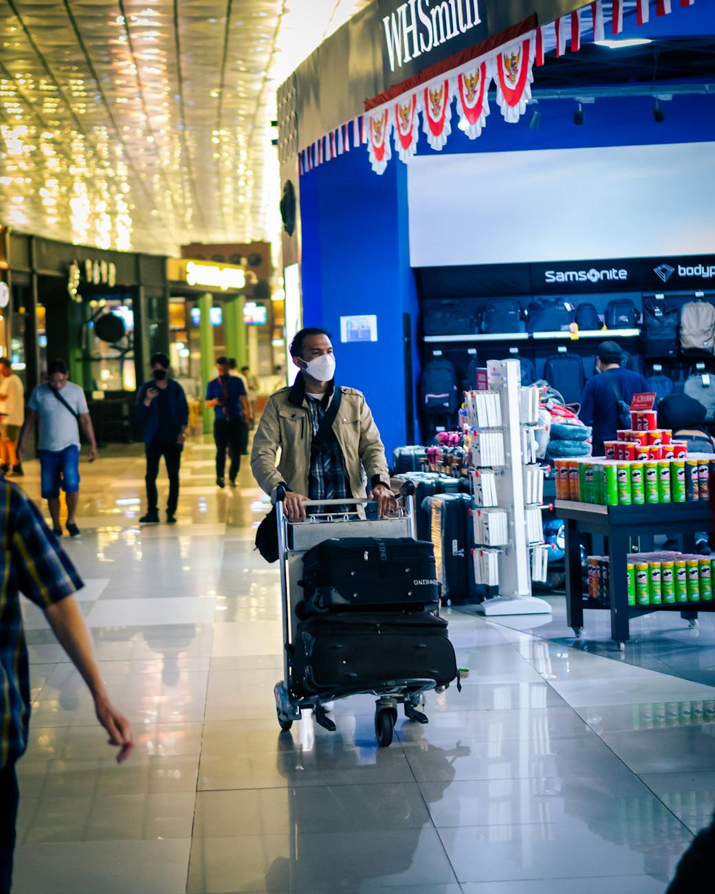 Man with luggage and mask in airport corridor near store. Busy day travel scene.