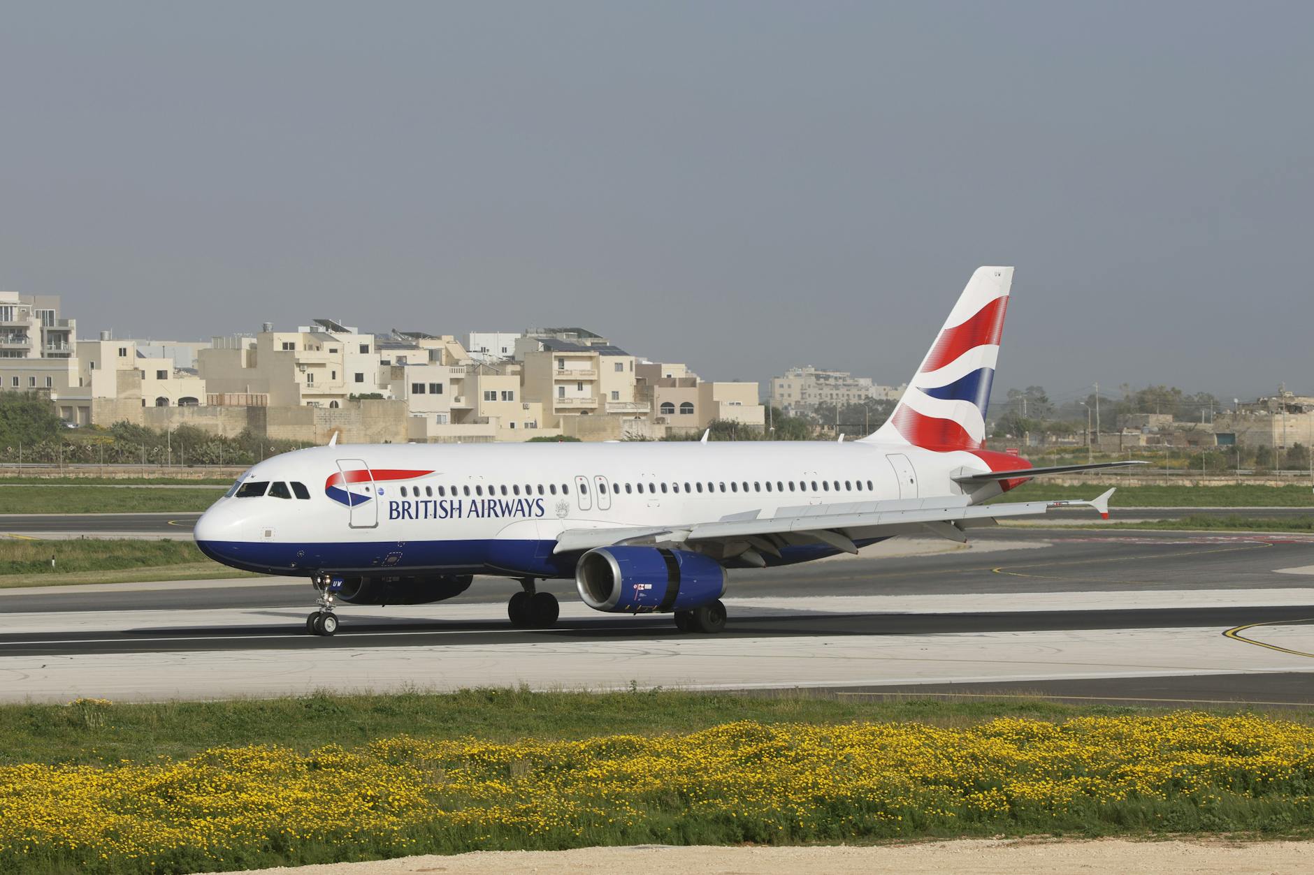 A British Airways Airbus A320 aircraft on a runway, with urban background.