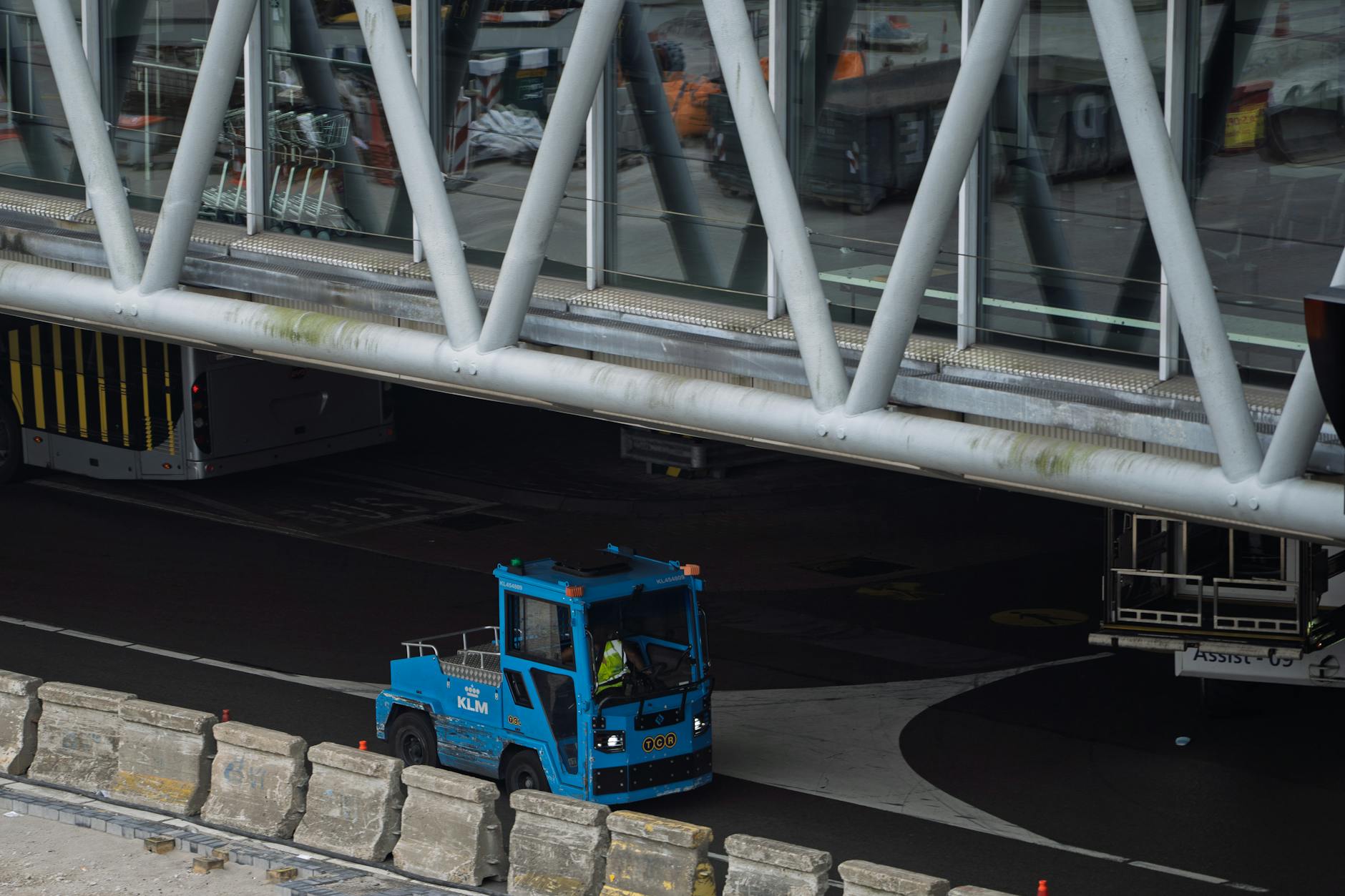 Blue airport ground vehicle moving under a jet bridge at a busy terminal.