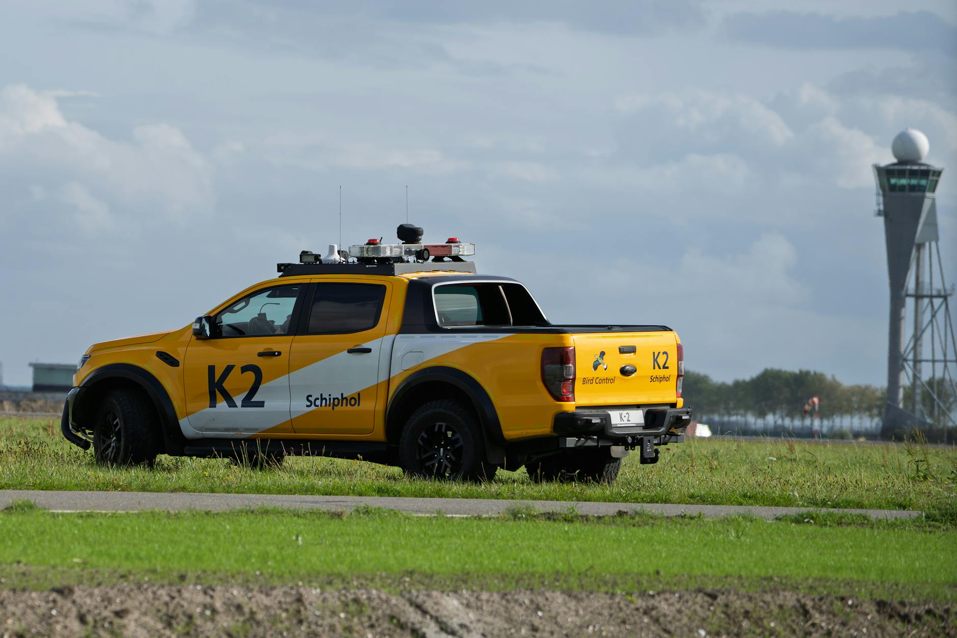 Schiphol Bird Control vehicle parked on grass near airport tower under cloudy skies.