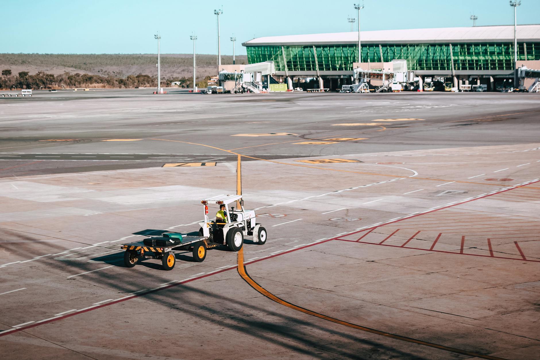 A luggage cart on the airport tarmac near a modern terminal building under clear skies.