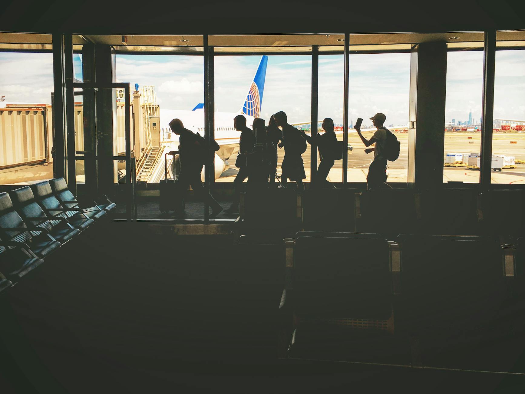 Silhouettes of travelers at an airport gate, airplane and city skyline visible through windows.