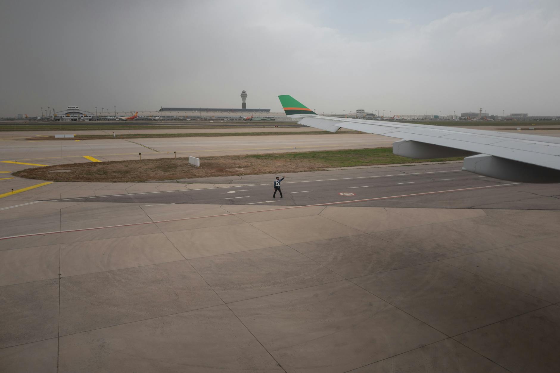 View of an airport runway with aircraft wing, control tower, and ground staff guiding on a cloudy day.