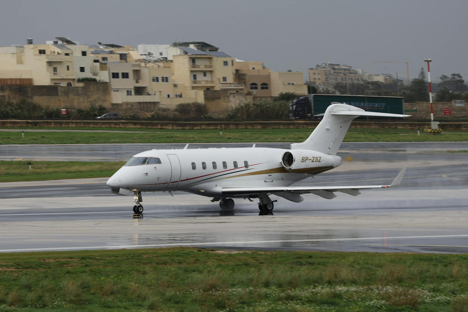 A private jet taxis on a wet runway at an airport with buildings in the background.