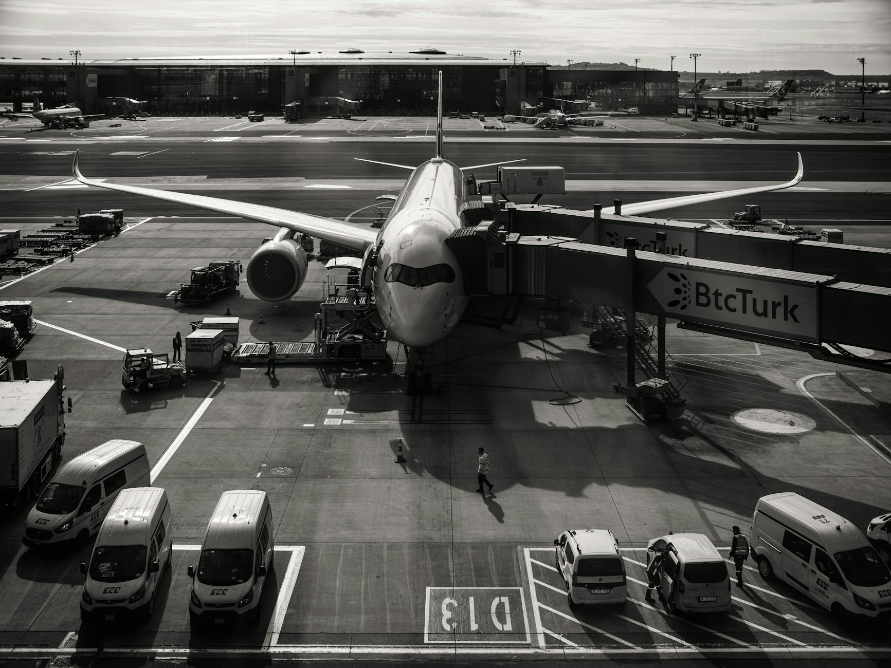 Monochrome image of airplane at airport gate with vehicles and jetway during the day.