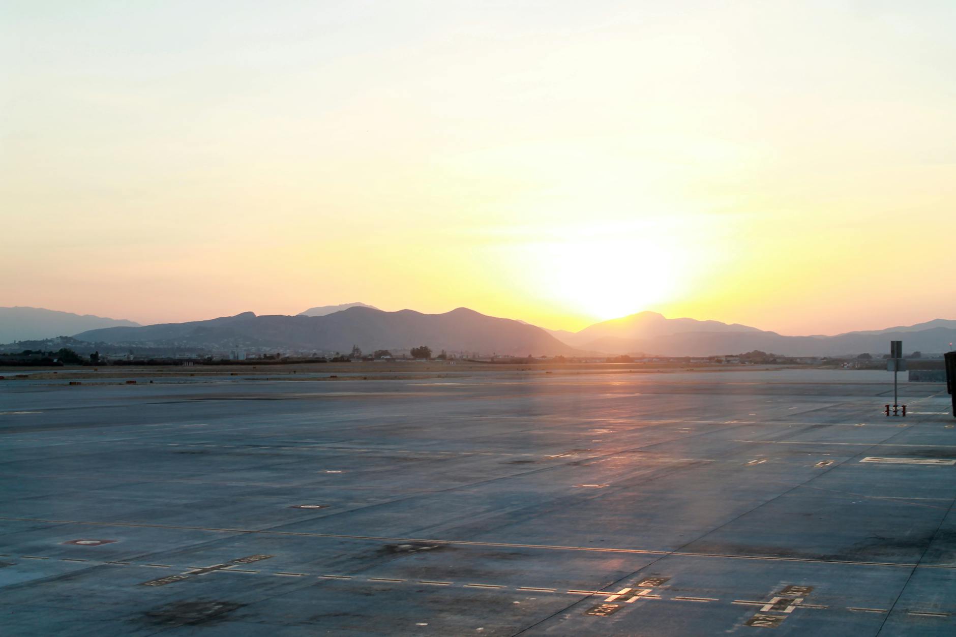Tranquil view of an airport runway with the sun rising over distant mountains, casting a gentle glow.