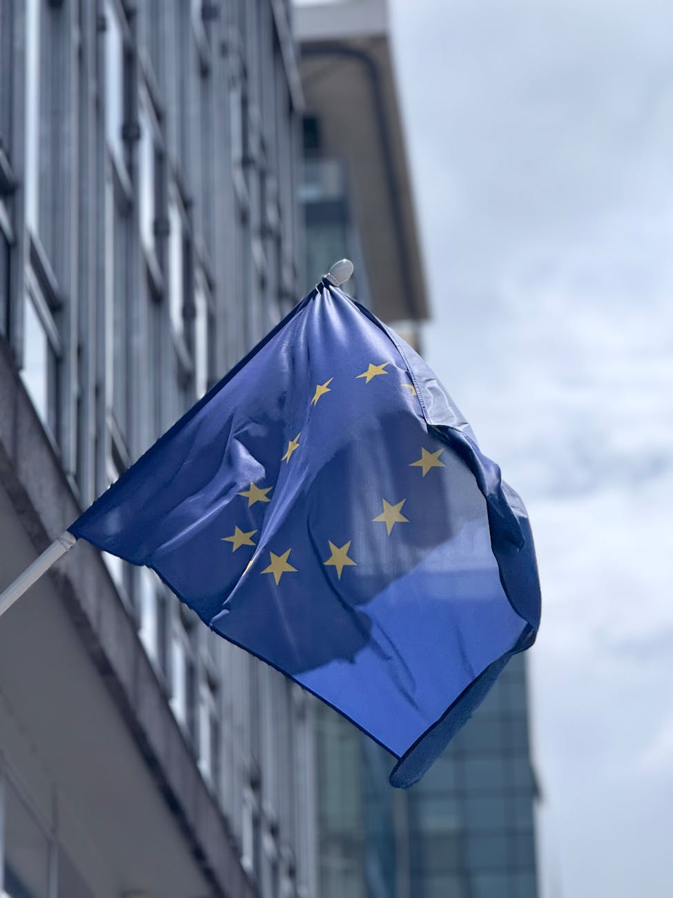 European Union flag waving in front of a modern glass building on a cloudy day.