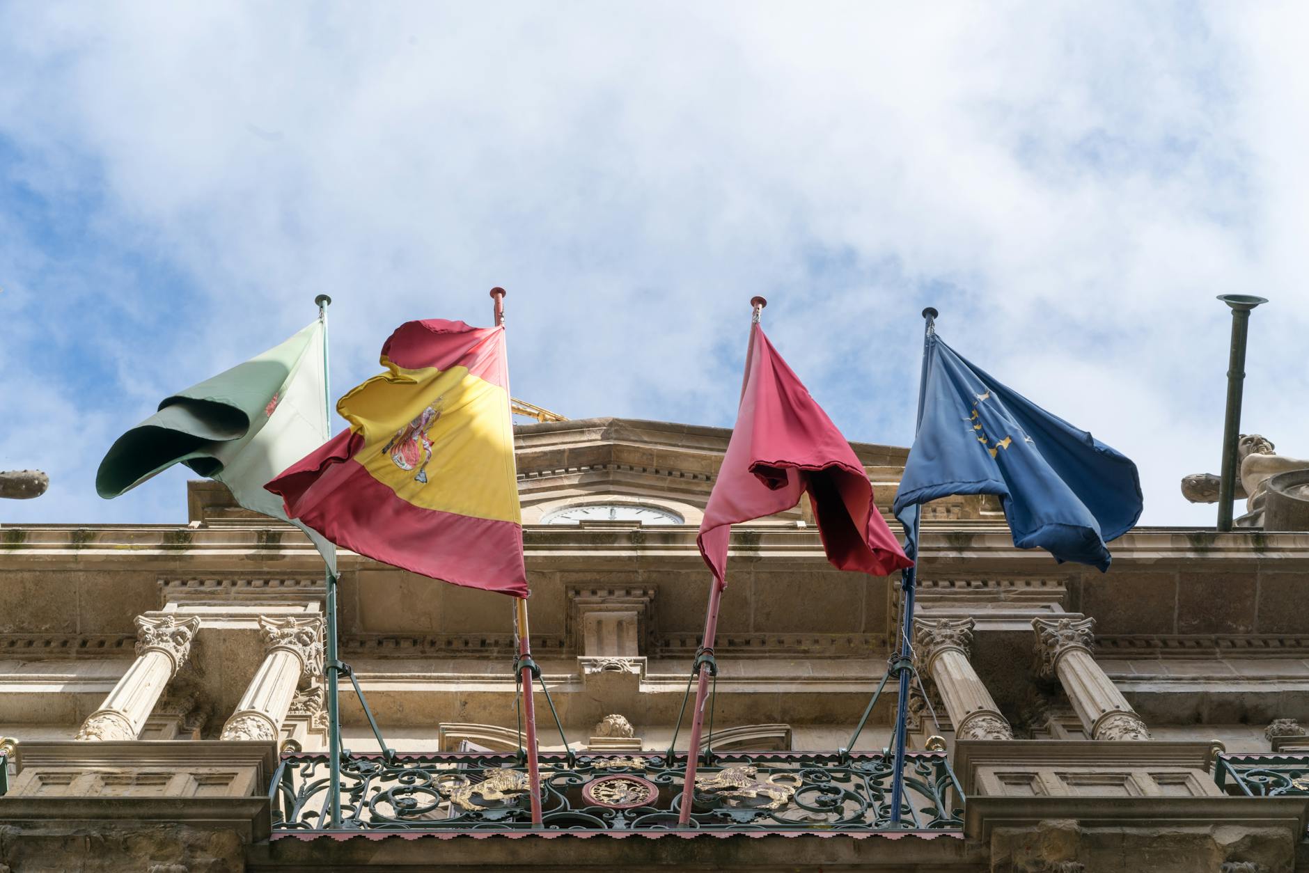 Flags of Spain, Italy, and EU on a historic building facade, symbolizing international unity.