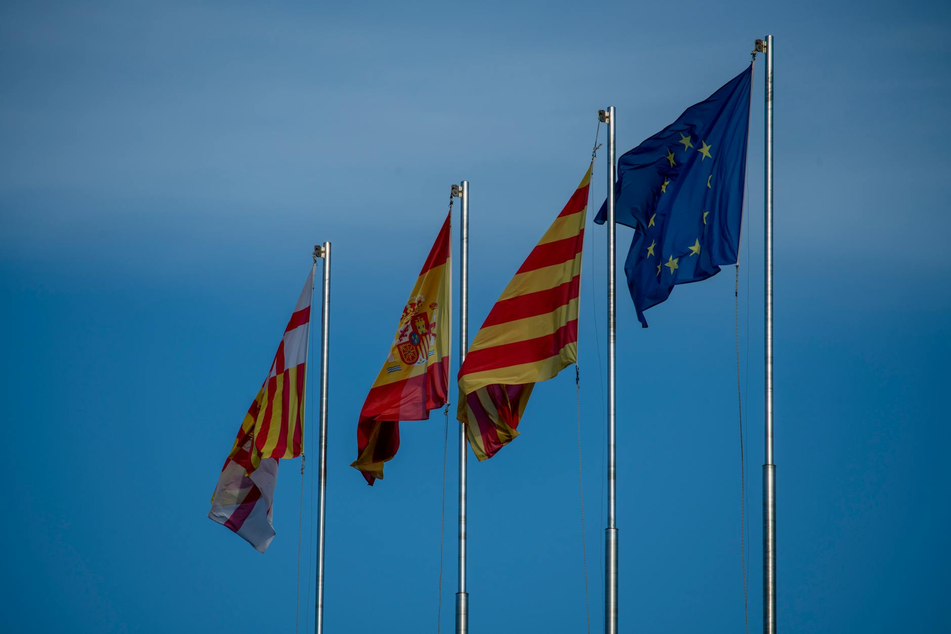 Flags of Spain, Catalonia, and Europe on flagpoles against a blue sky in Barcelona.