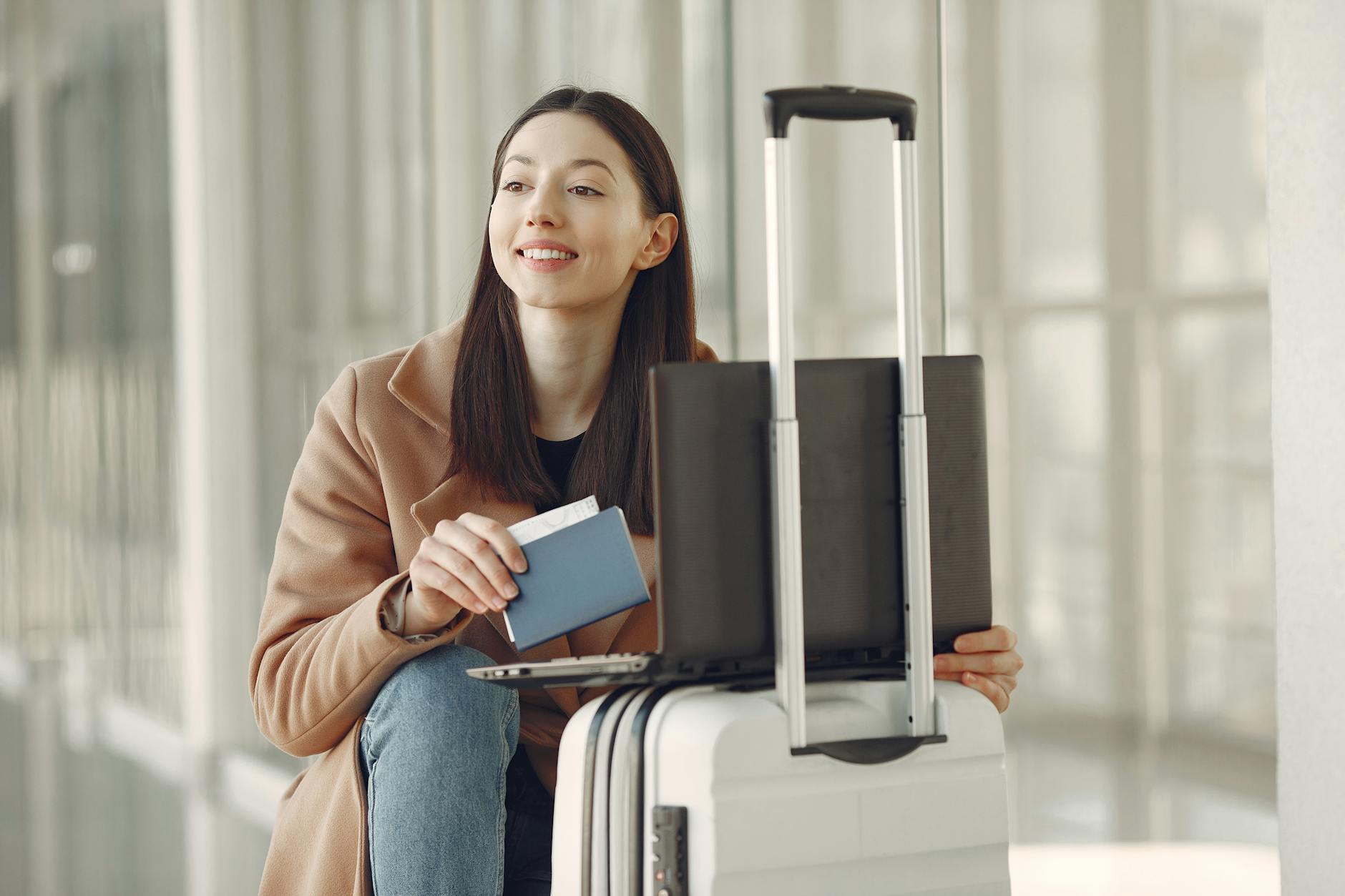 A woman sitting with her laptop and luggage, holding a passport, waiting in an airport terminal.