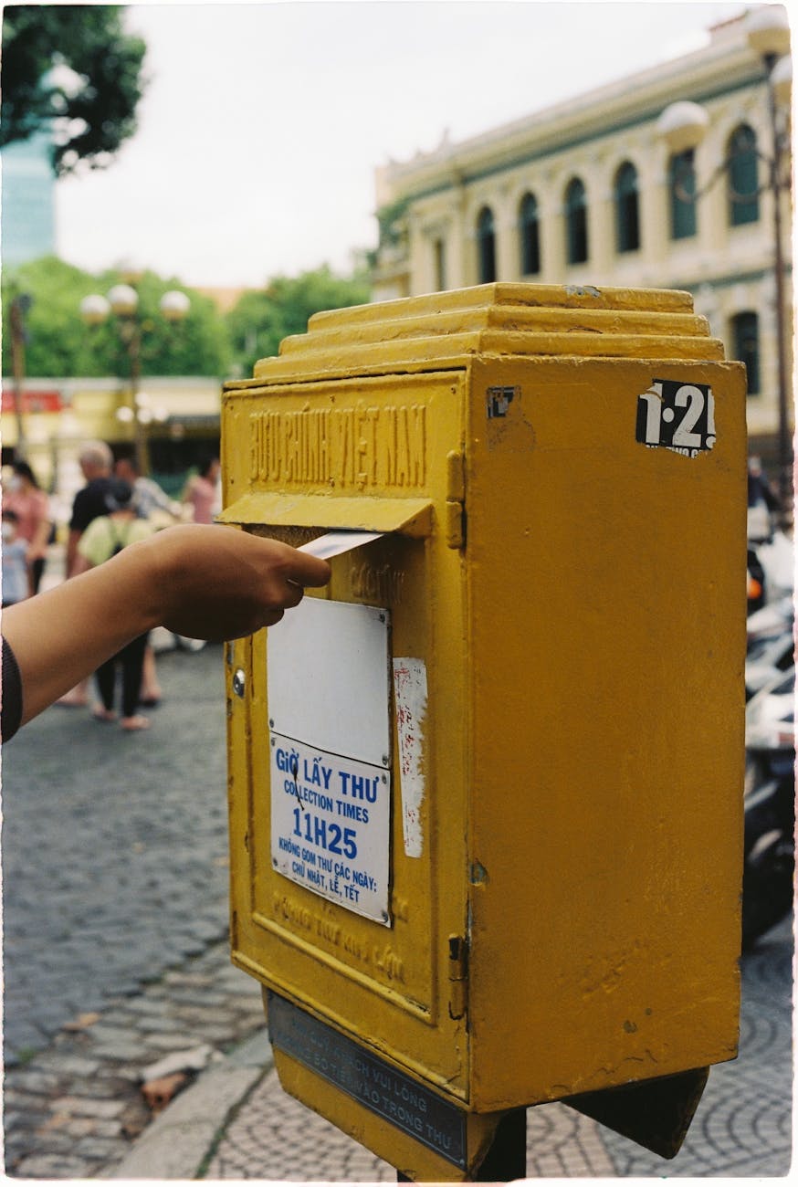 A hand posting a letter into a yellow mailbox on a busy Vietnamese street.