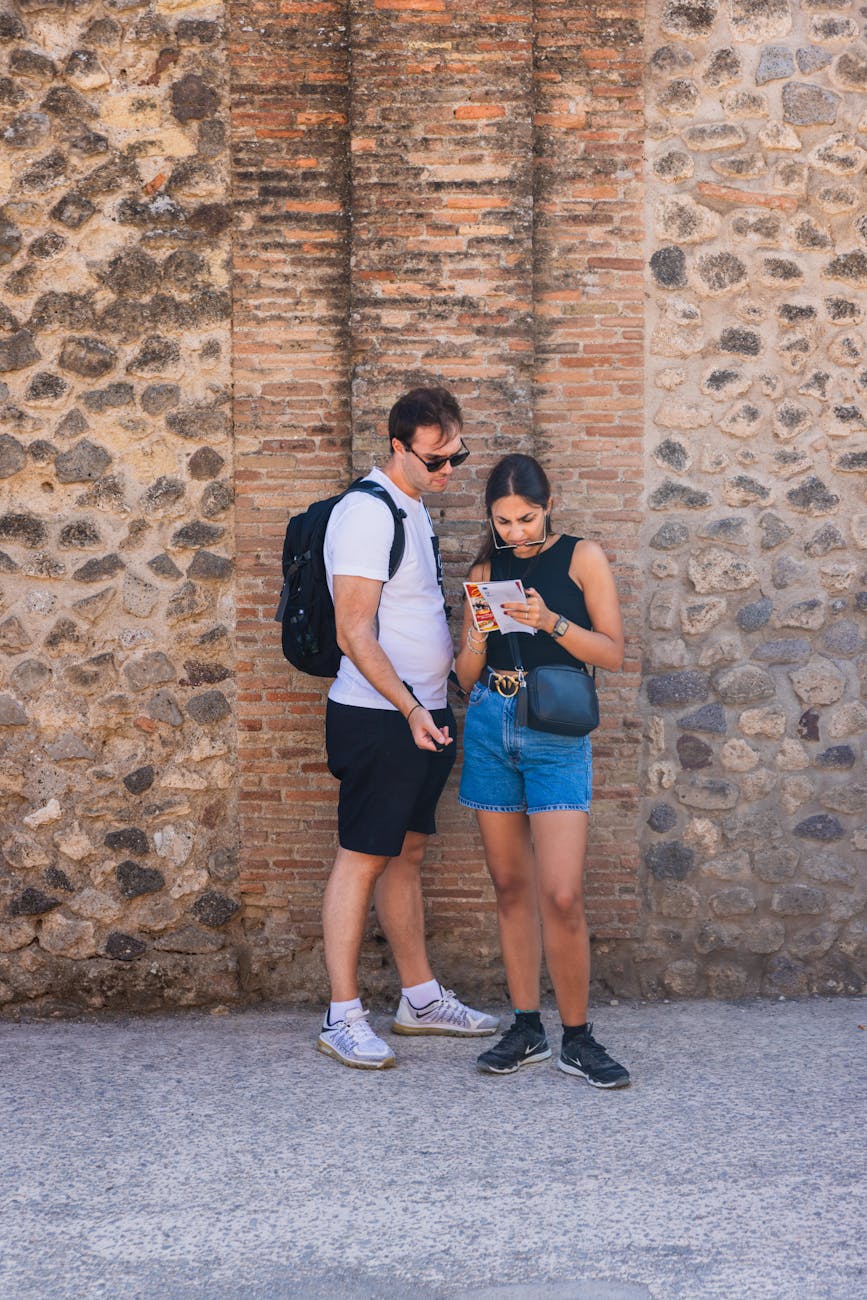 A couple stands against a stone wall in an old town, studying a map for directions.