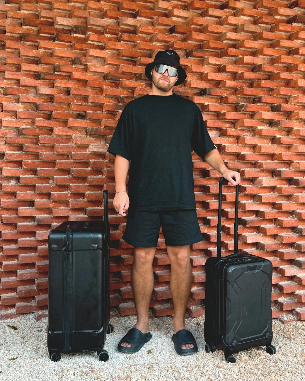Man in black attire with suitcases stands against a red brick wall in Mexico.