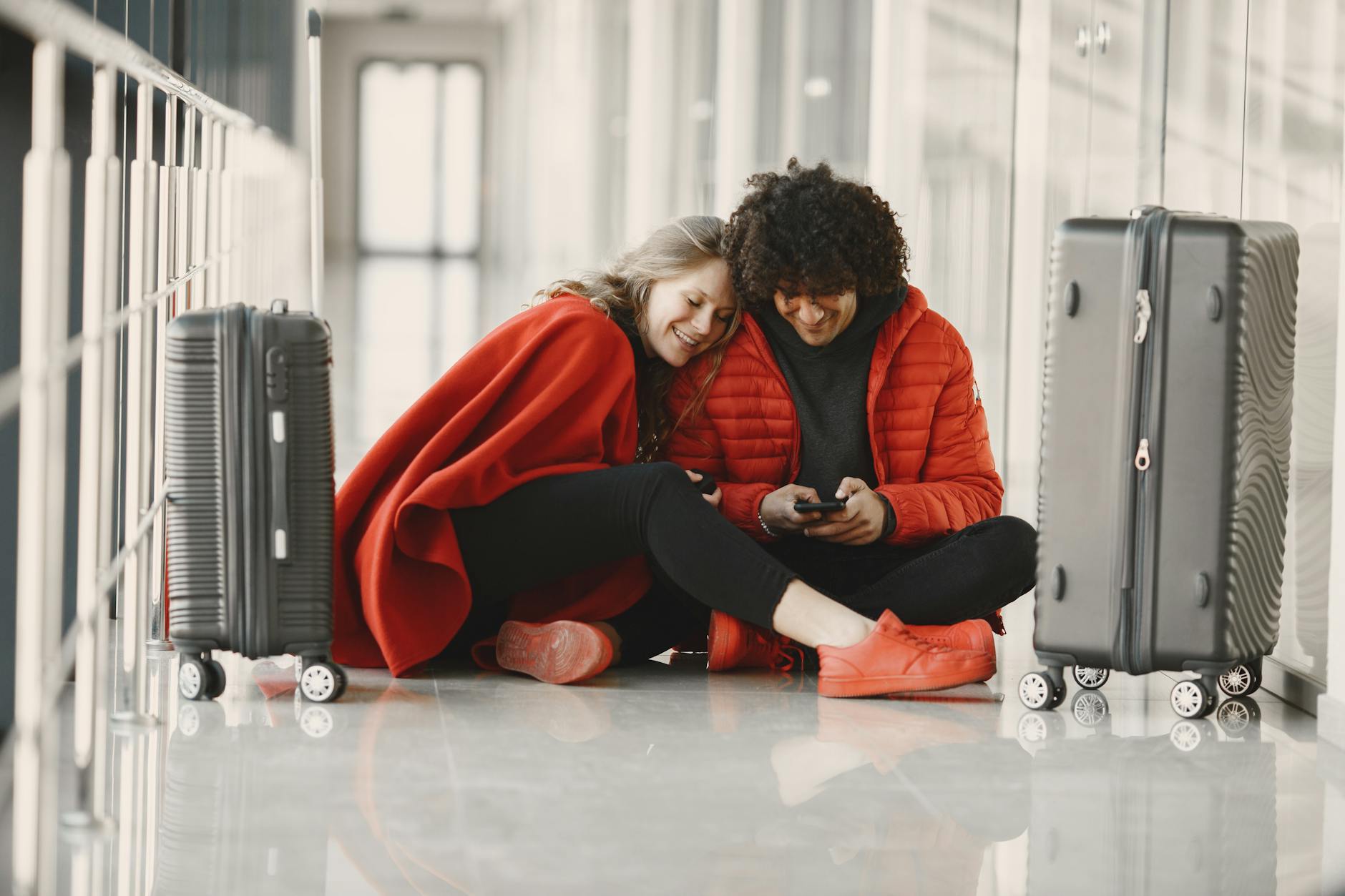 Happy couple sitting on the airport floor with luggage, sharing a moment using smartphone.