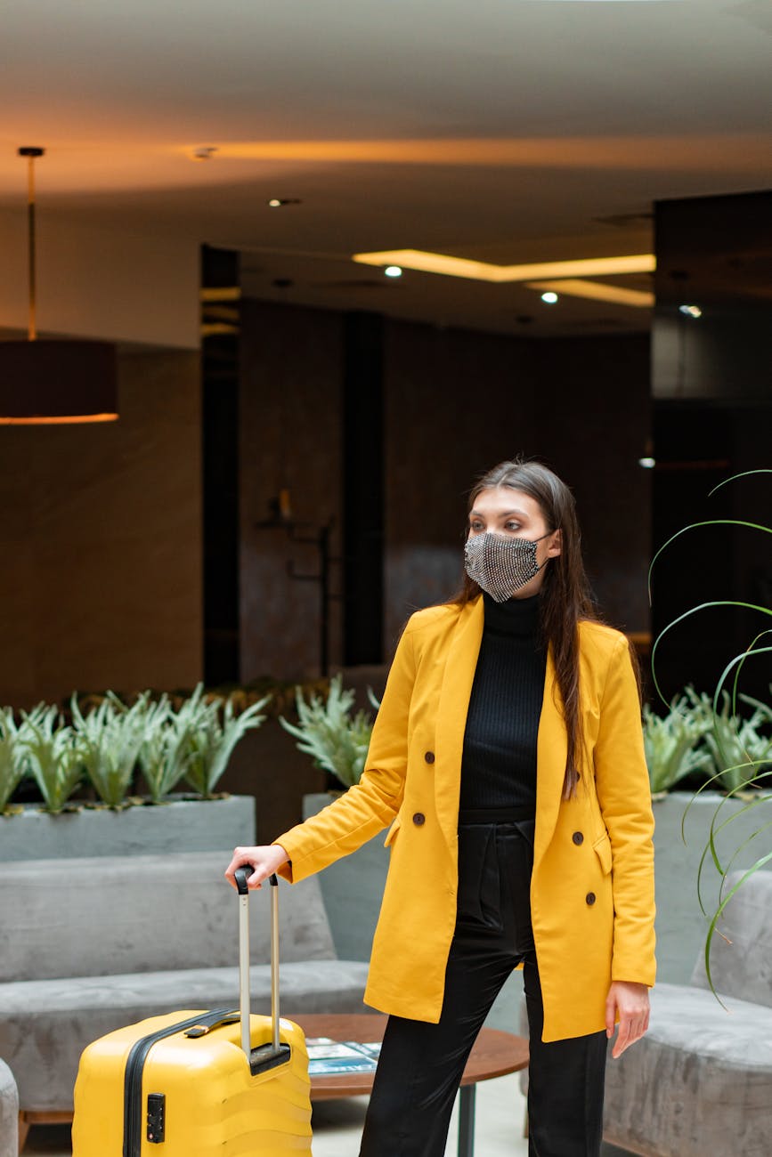 Fashionable woman with luggage in hotel lobby wearing mask, representing travel safety.
